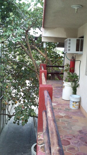A covered outdoor area with a stone floor, a red barrier, a white air conditioning unit, and a potted plant.