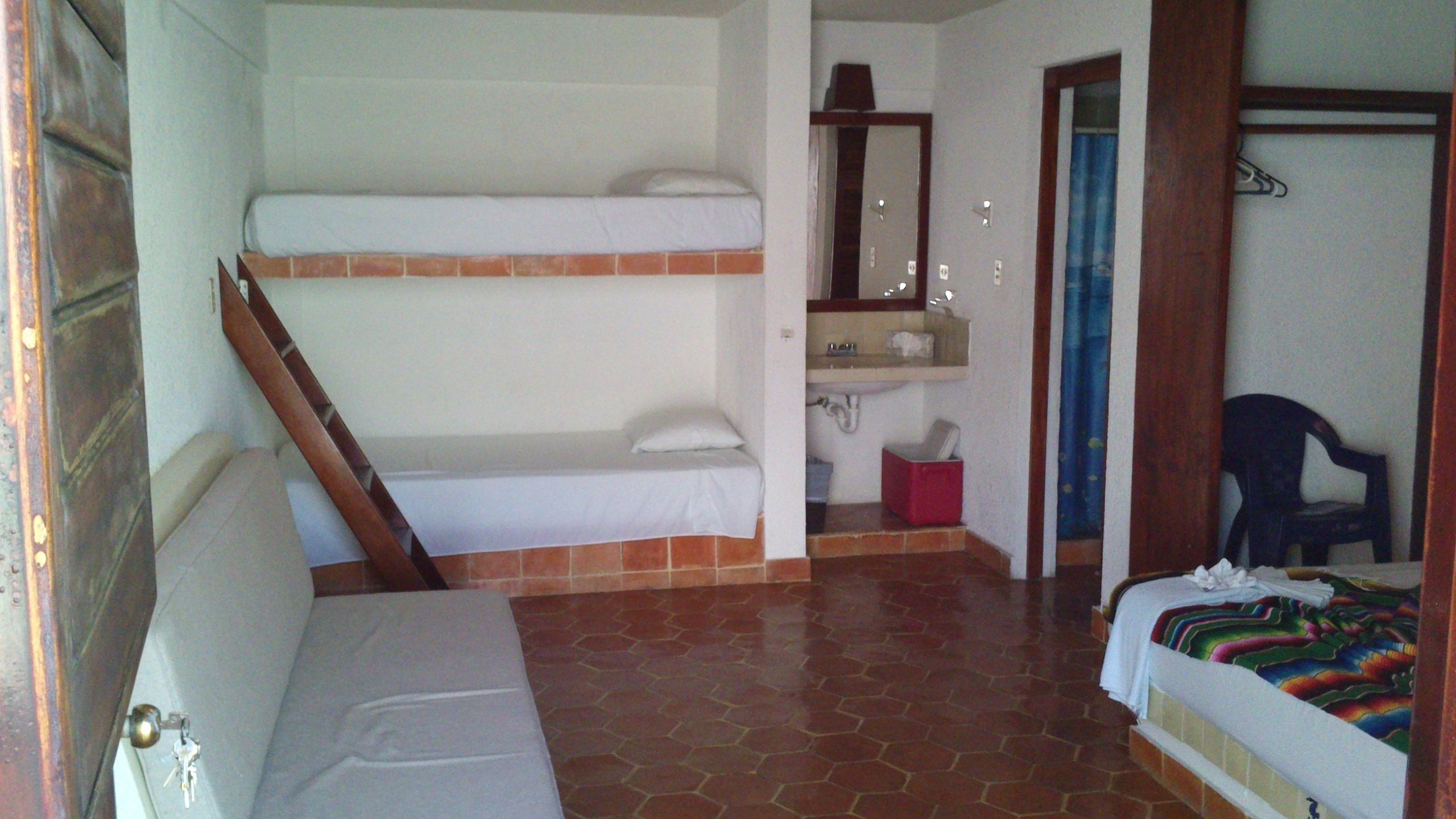 A room with a bunk bed, a small sofa, a bathroom with a sink and mirror, and a chair.