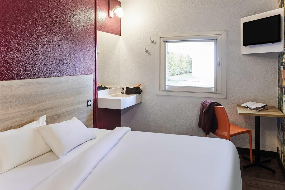 A hotel room with a bed, a desk, a chair, a window, and a TV mounted on the wall.