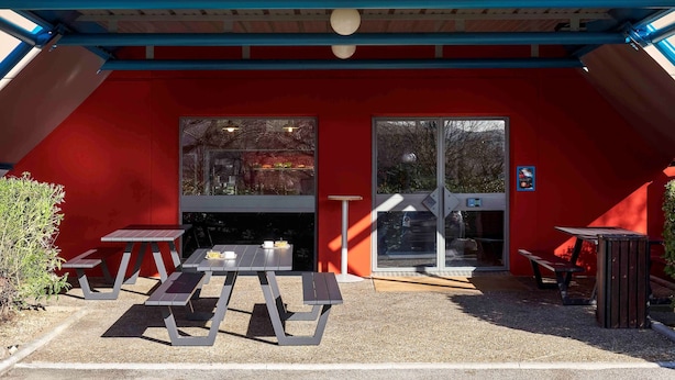 L’entrée d’un hôtel avec une façade rouge, une enseigne jaune au-dessus de la porte et un espace aménagé avec des tables de pique-nique et des bancs.