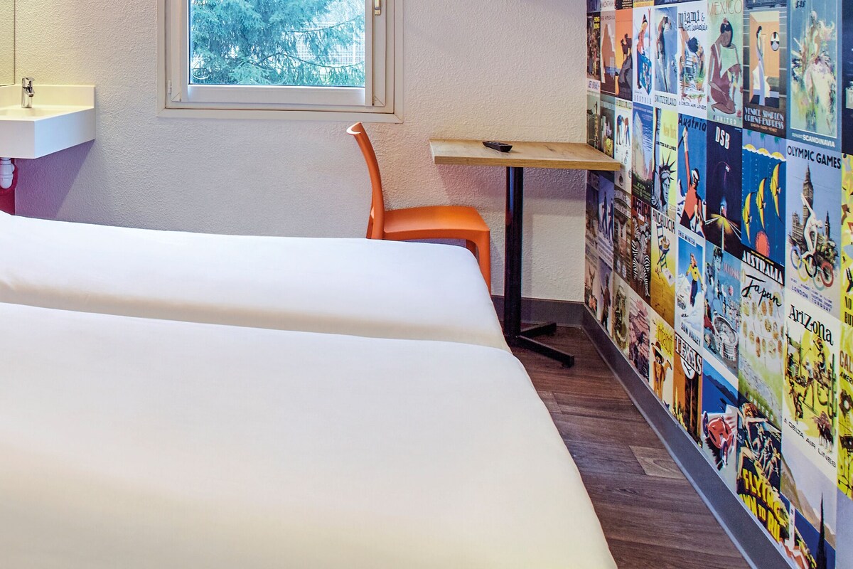 Une chambre d’hôtel avec une tête de lit en bois, un lit blanc, un petit lavabo, une chaise orange et un mur décoré d’affiches de voyage.