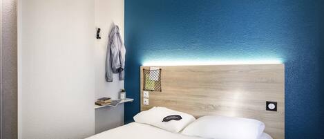 Habitación de hotel con cama, cabecera con luz, repisa con un libro y una planta, y pared con detalle azul.