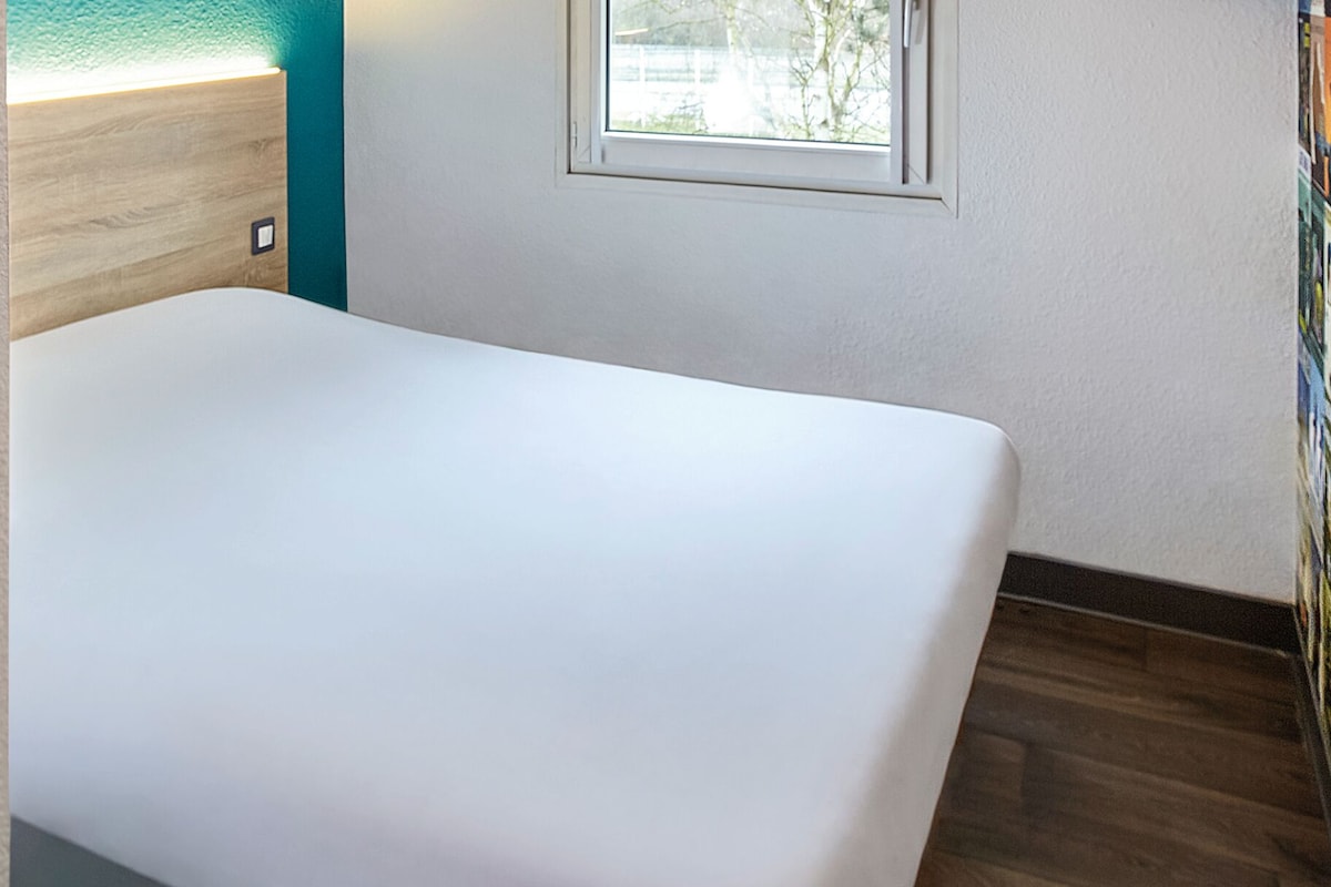 Une pièce avec un lit blanc, une tête de lit en bois, une petite fenêtre et une porte.