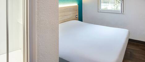 A room with a white bed, a wooden headboard, a small window, and a door.