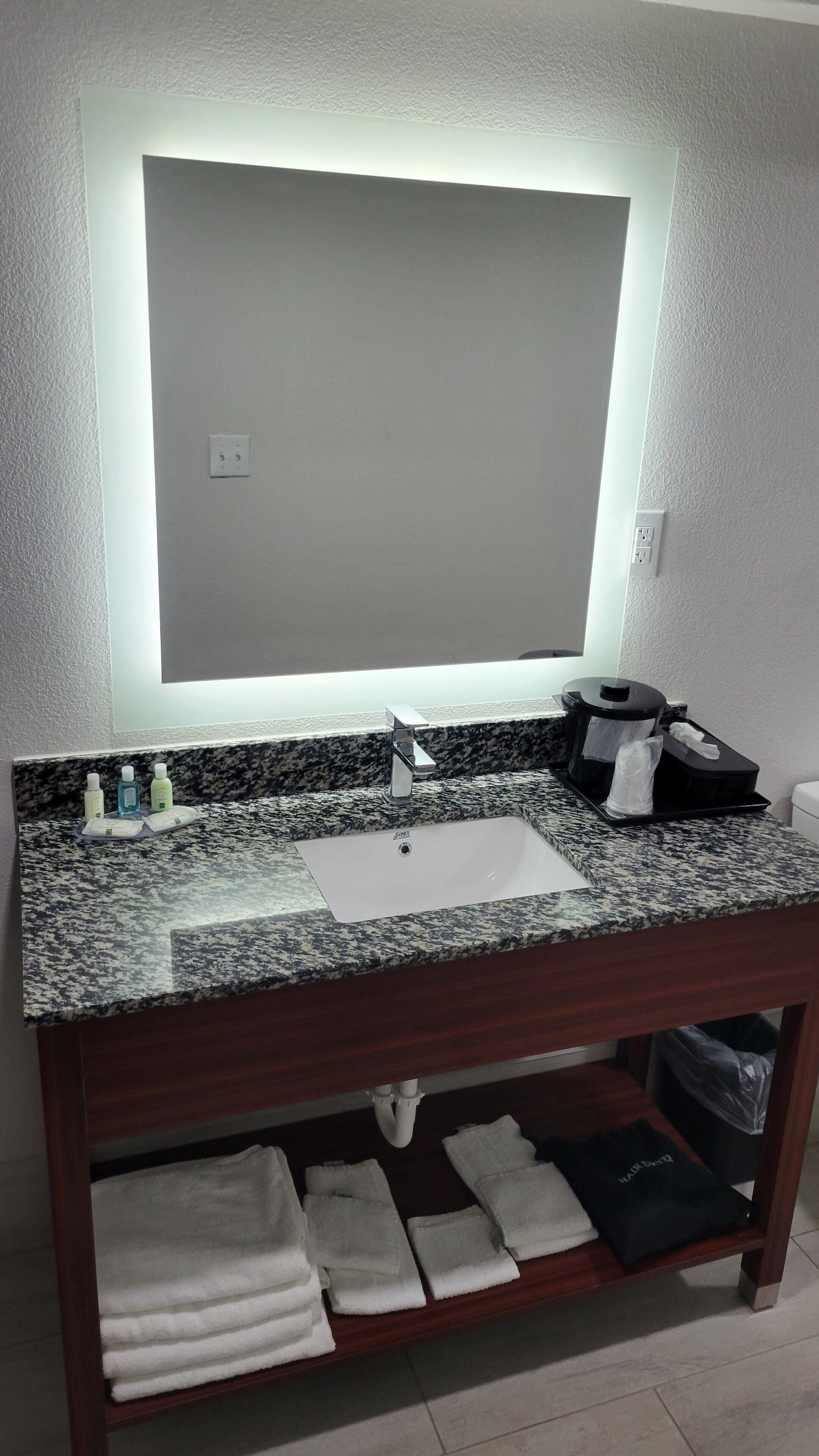 Un baño con un espejo grande, un lavabo y un mueble de tocador con encimera de granito. Hay toallas y artículos de aseo en el mueble.