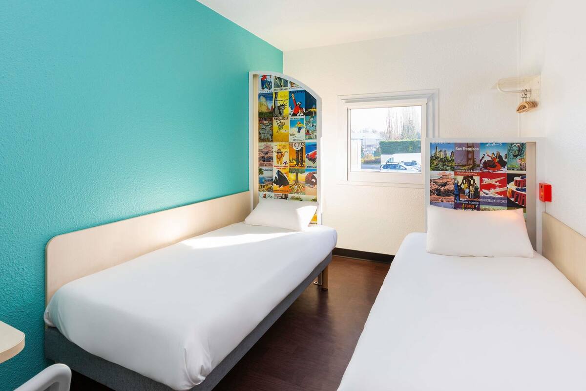 Habitación de hotel con dos camas individuales, una mesita pequeña, una ventana con vistas y una pared decorada con carteles de viajes.