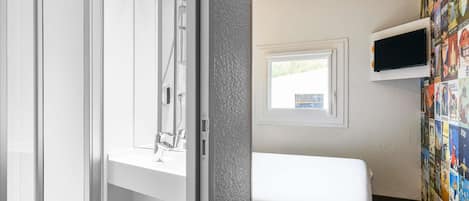 A room with a bed, a TV mounted on the wall, a window, and a bathroom with a shower.
