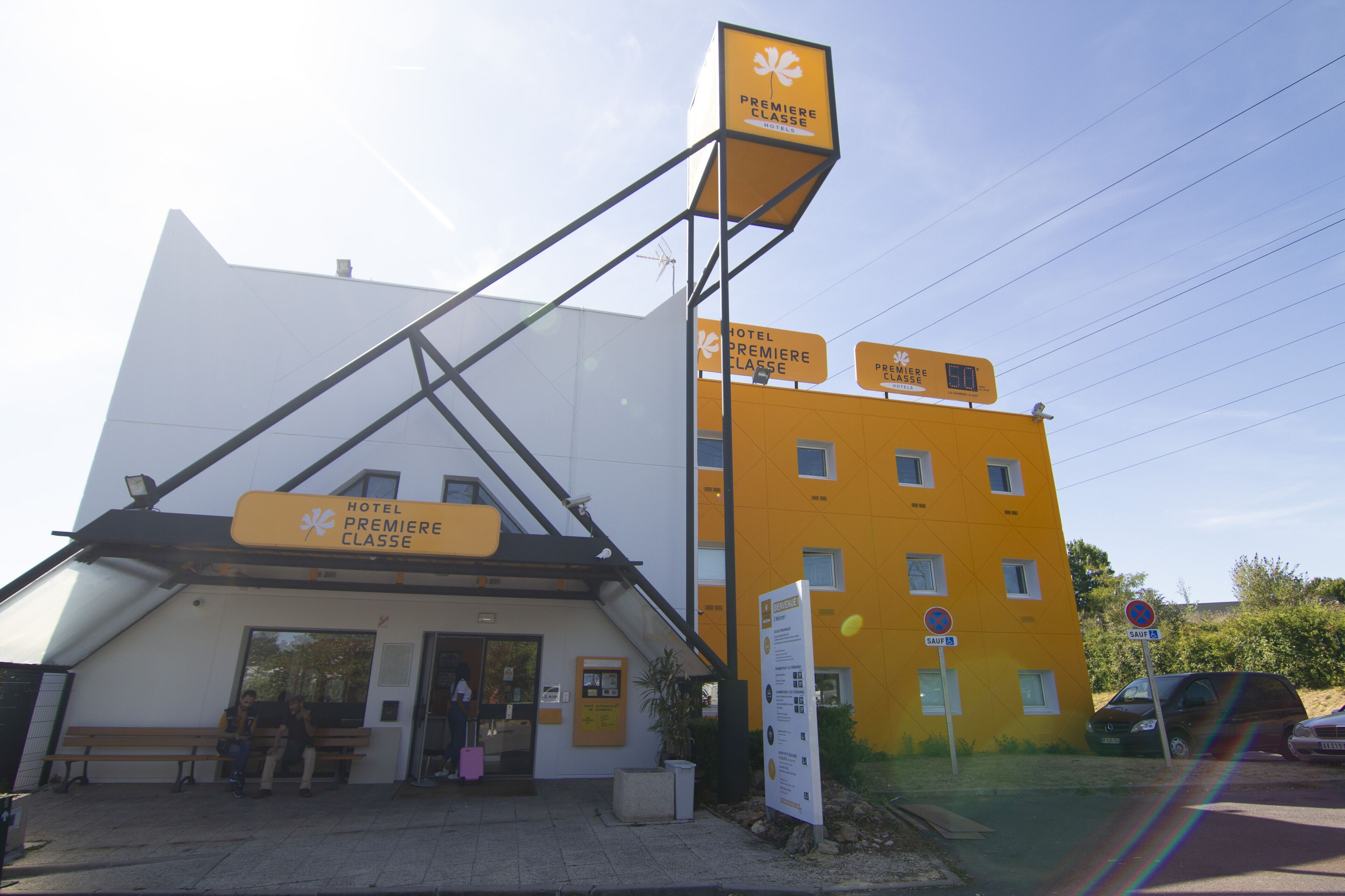 A hotel with a prominent yellow and white facade, a sign indicating 'HOTEL PREMIERE CLASSE', and a visible entrance with a bench outside.