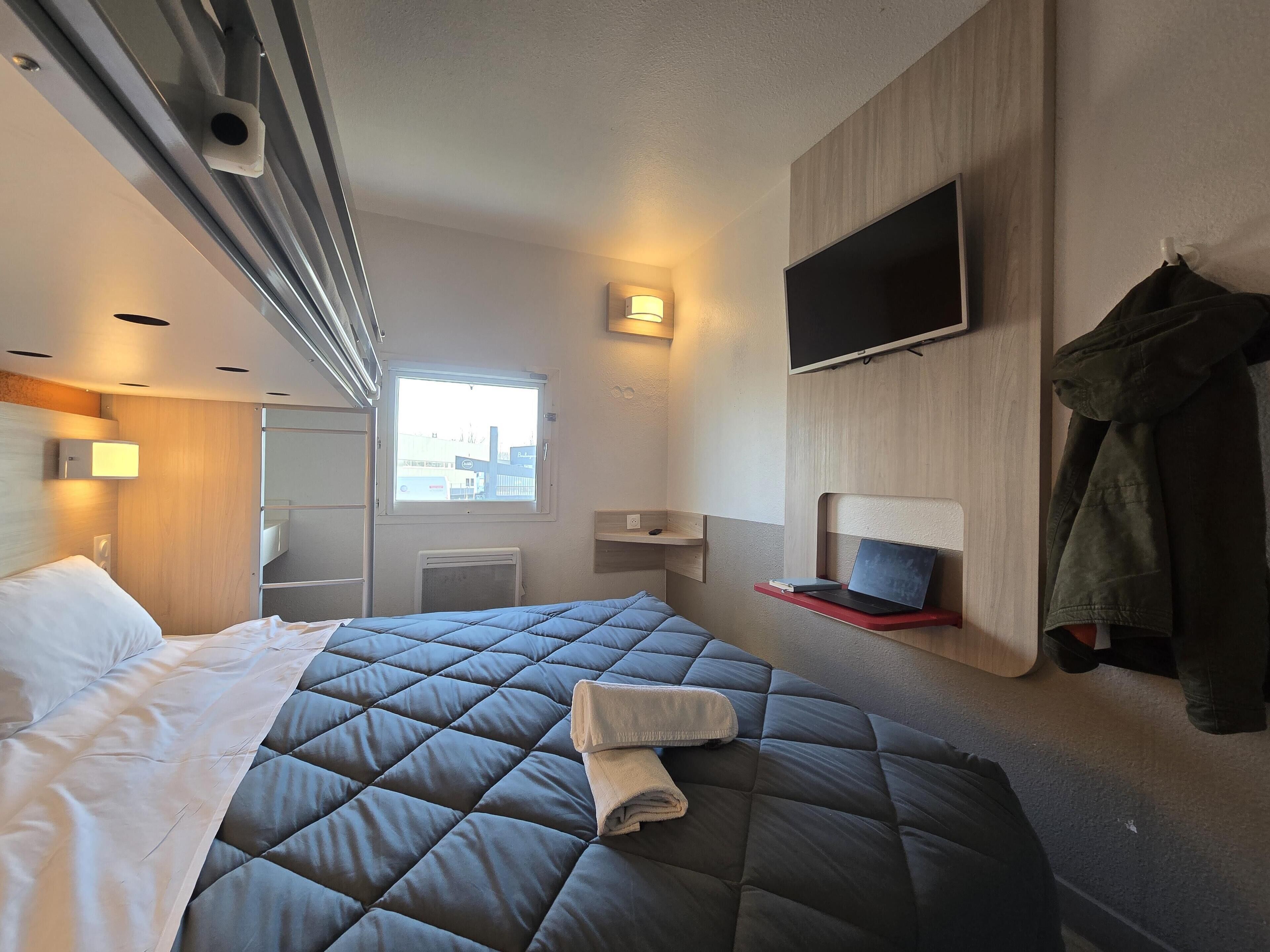 A modern hotel room with a large bed, a flat-screen TV mounted on the wall, and a window offering a view of the city.