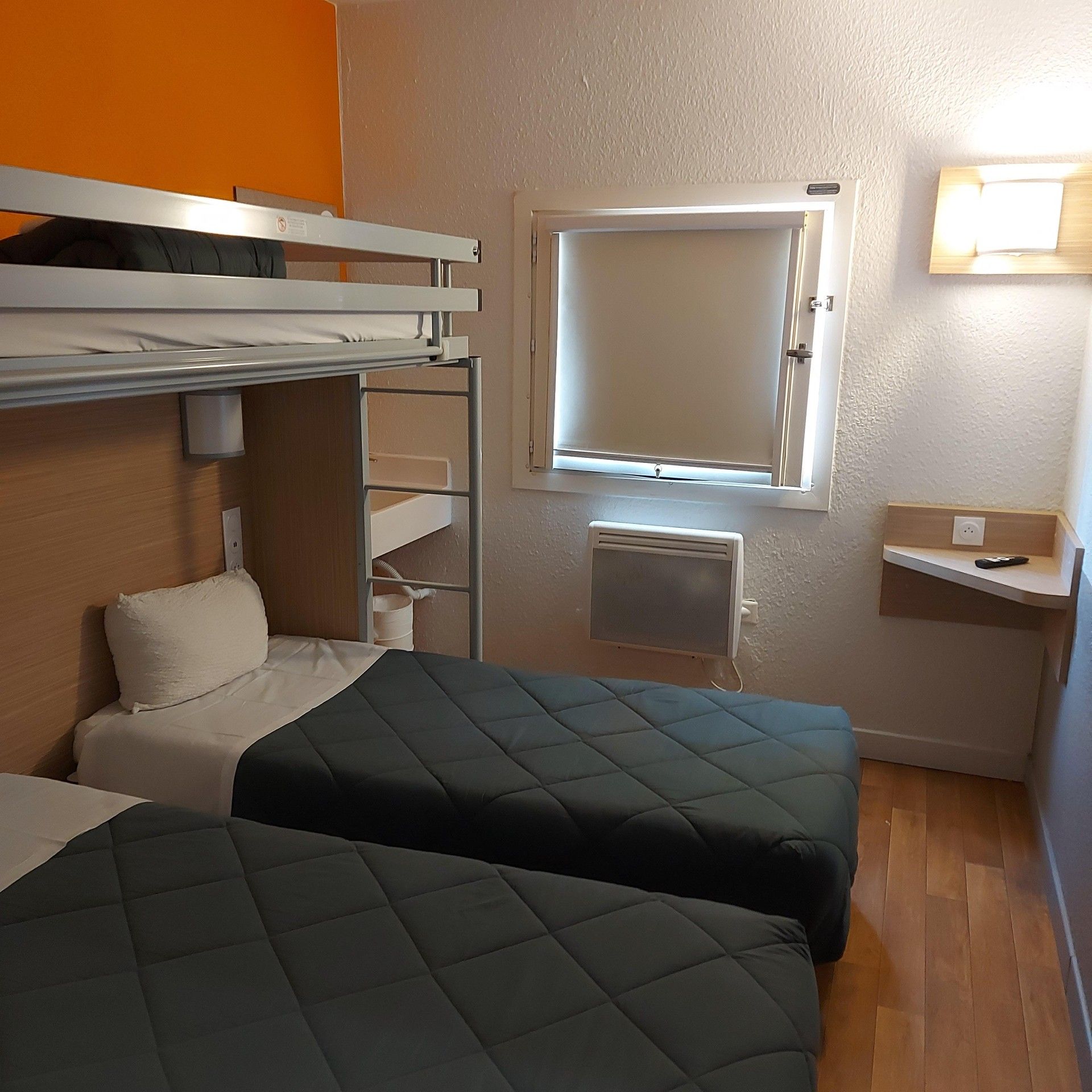 A room with bunk beds, a small window, a heater, and a shelf with a remote control.