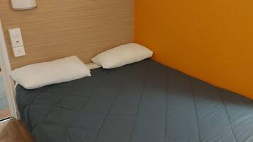 A single bed with a grey quilt, two white pillows, a wooden headboard, and a wall with an orange accent.