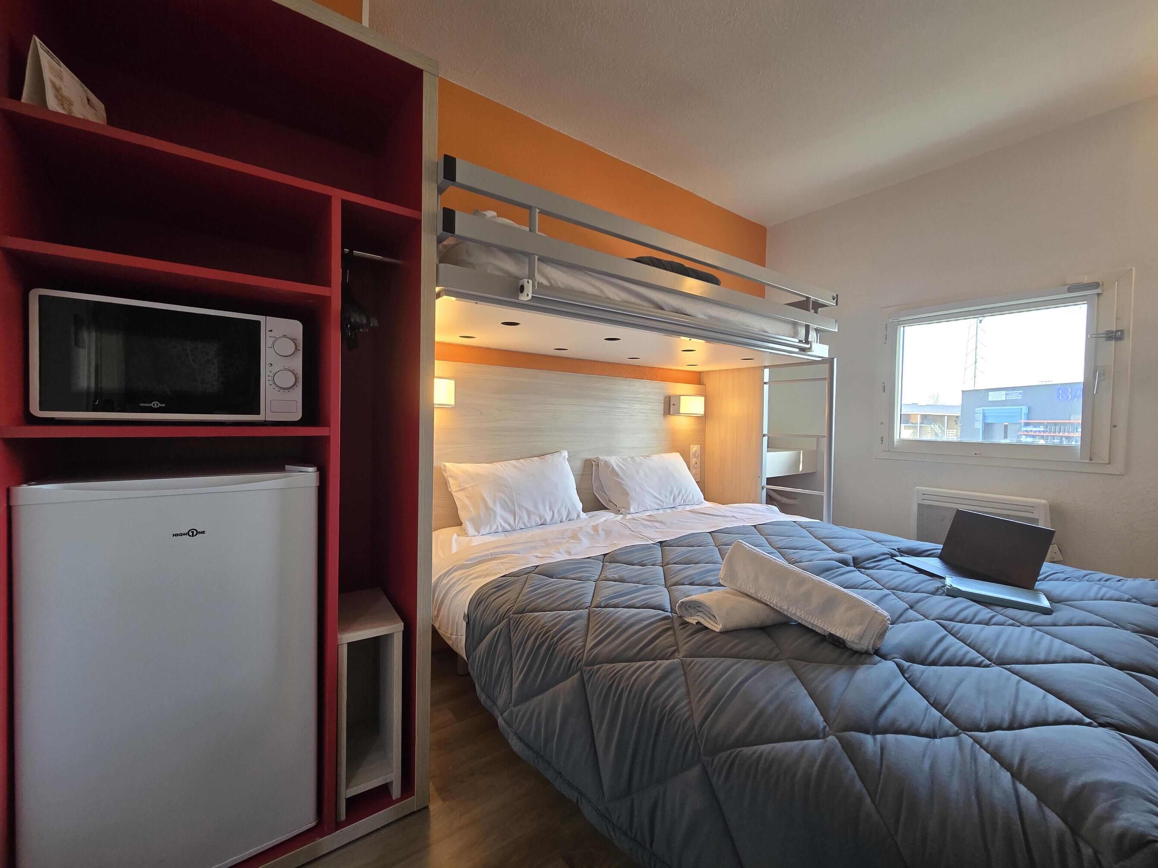 Ein kleines Hotelzimmer mit Bett, Mikrowelle, Minikühlschrank, Fenster mit Blick und integriertem Schreibtisch mit Laptop.
