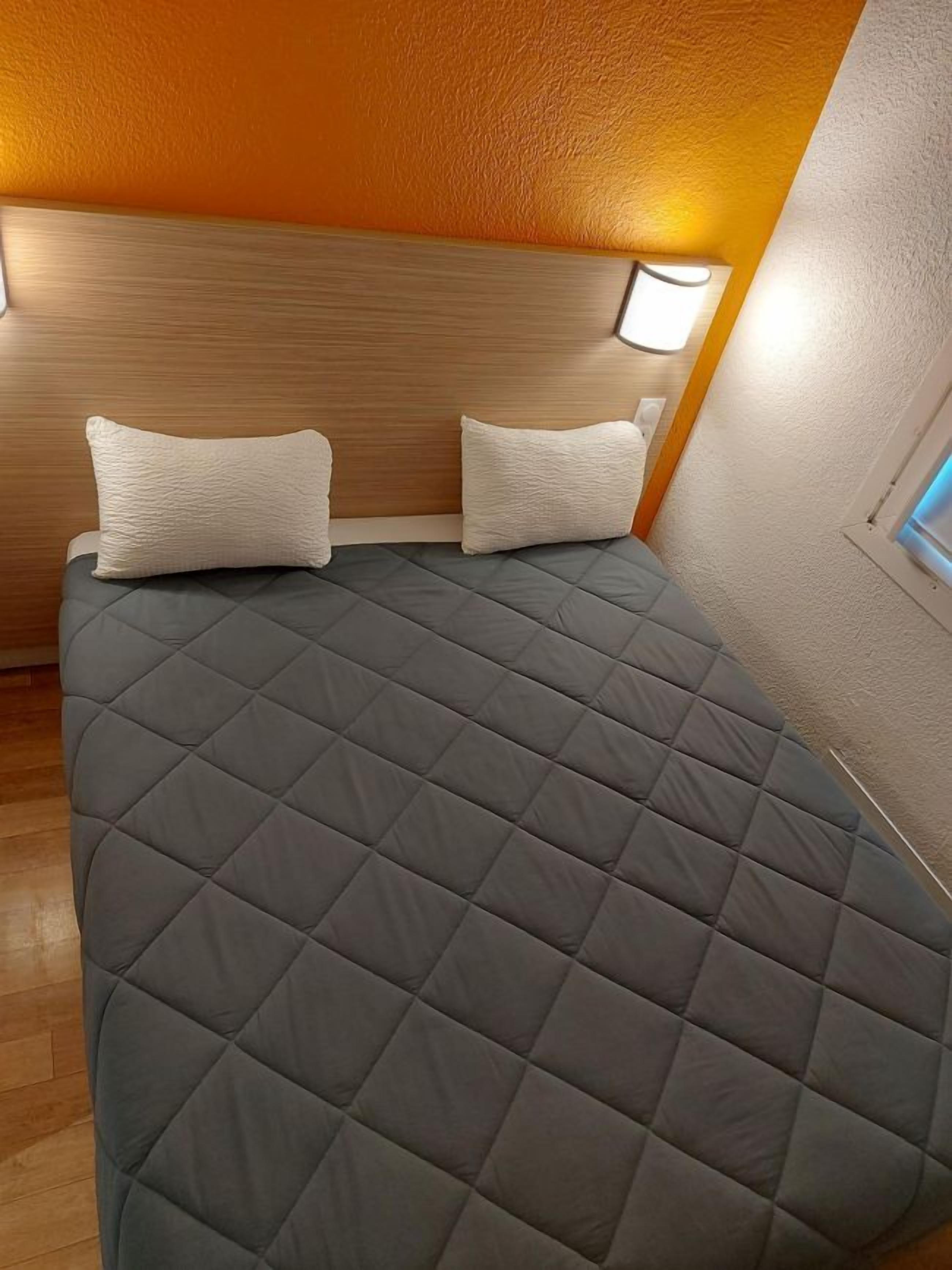A bed with a quilted grey cover, two pillows, and a headboard with a light fixture.