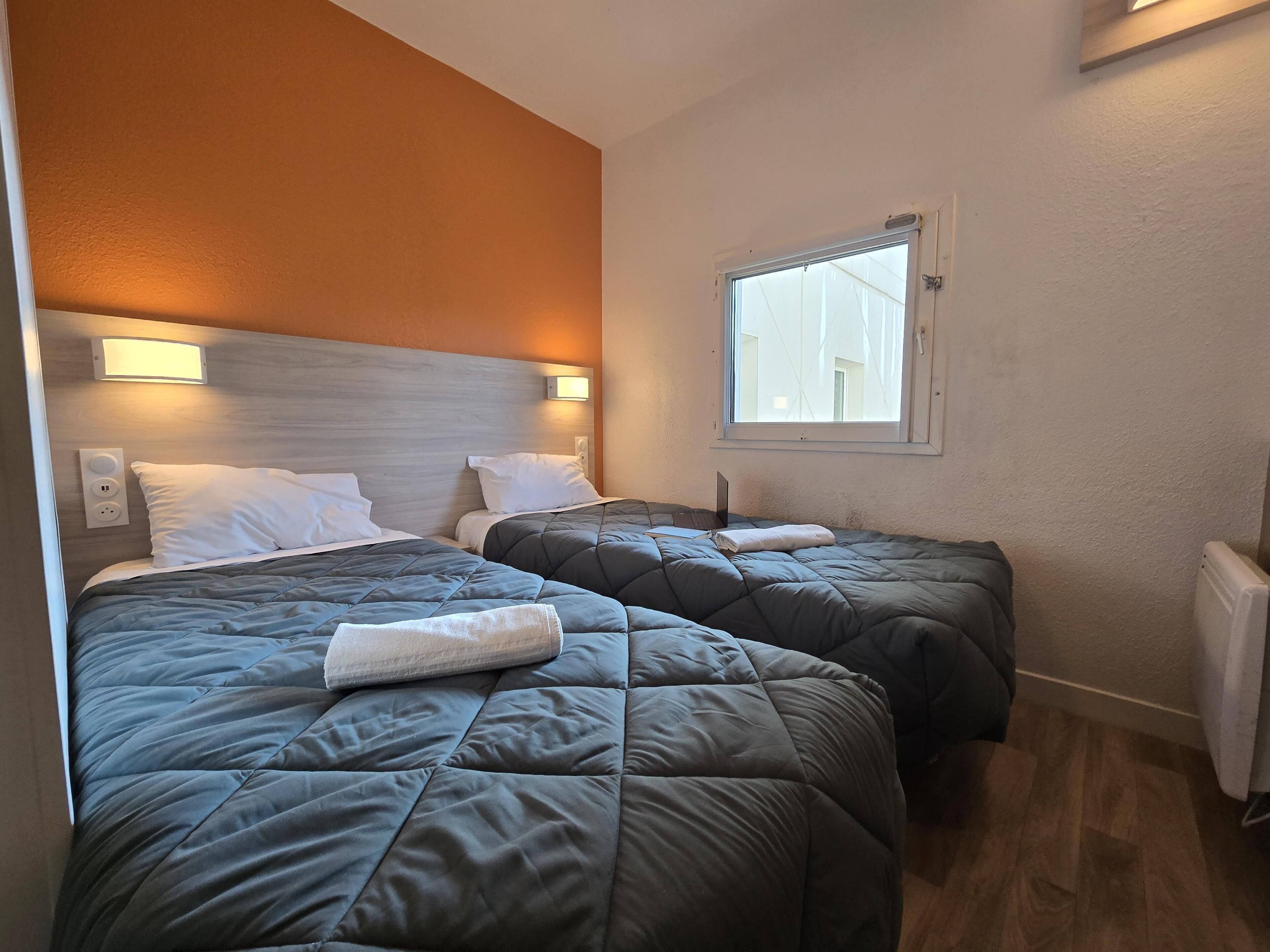 Une chambre d’hôtel avec deux lits, chacun recouvert d’une couette matelassée grise et agrémenté d’oreillers blancs. Une fenêtre offre une vue sur les bâtiments environnants, et un luminaire mural est fixé au-dessus des lits.