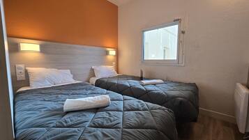 Une chambre d’hôtel avec deux lits, chacun recouvert d’une couette matelassée grise et agrémenté d’oreillers blancs. Une fenêtre offre une vue sur les bâtiments environnants, et un luminaire mural est fixé au-dessus des lits.