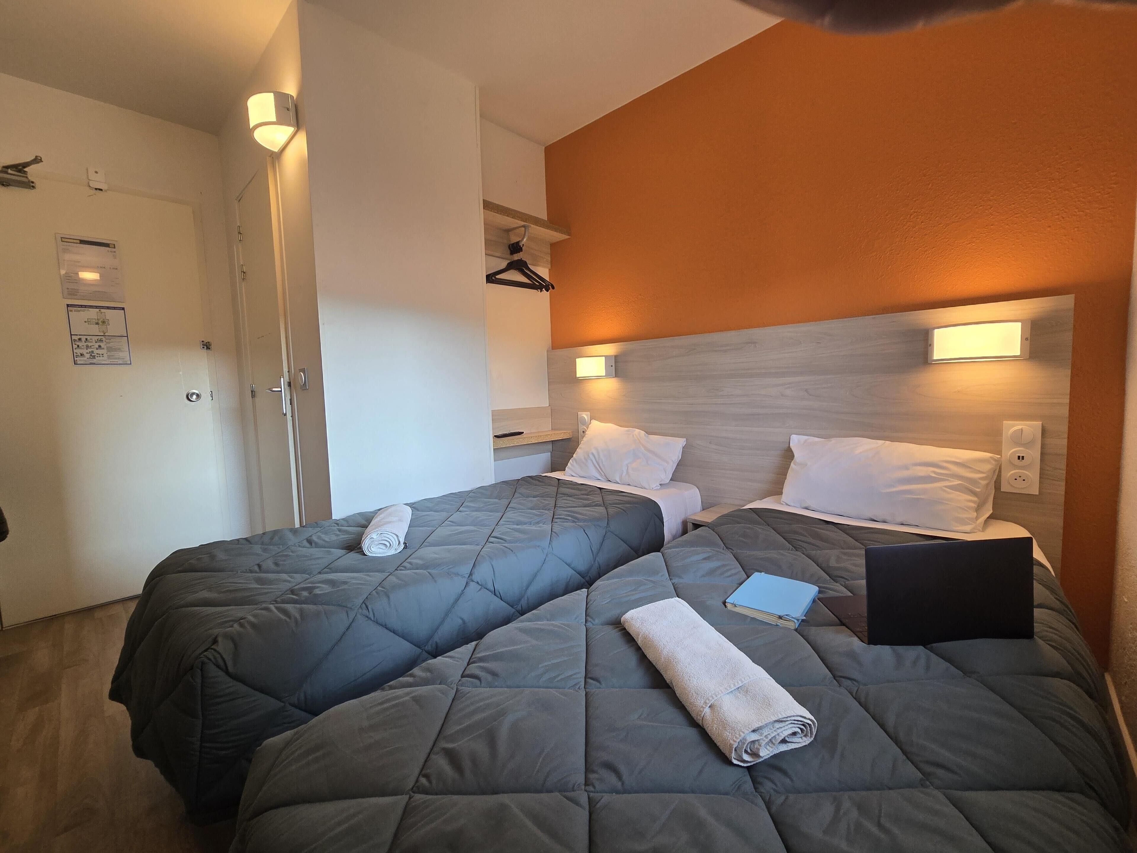 Habitación de hotel con dos camas, cada una con edredón acolchado gris y almohadas blancas. Hay una toalla en el suelo, un libro en una cama y un enchufe en la pared.