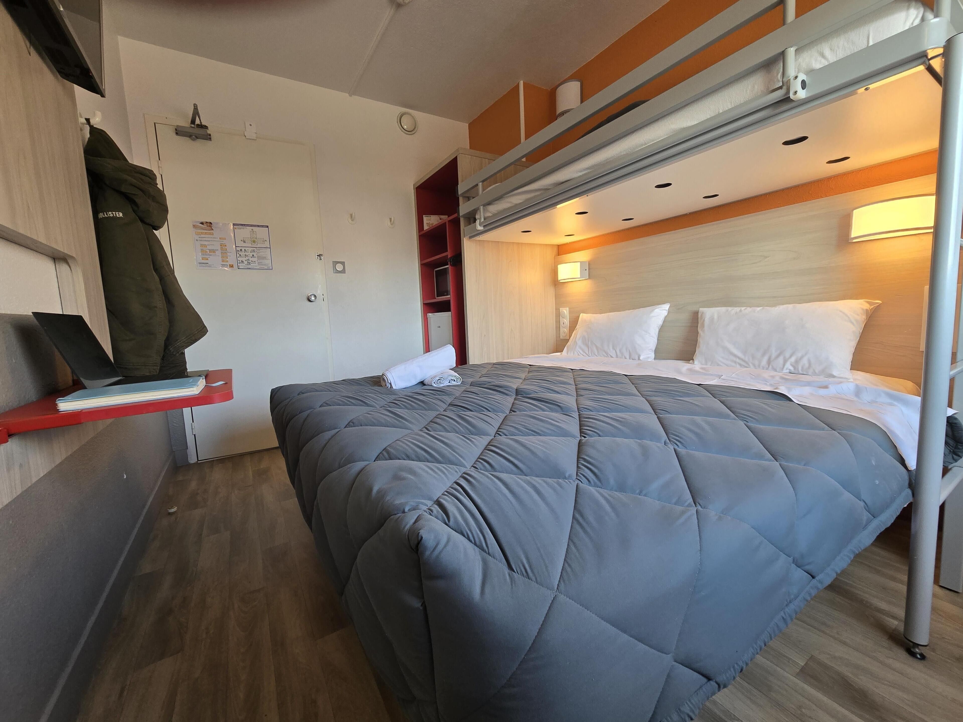 A compact hotel room with a bunk bed, a desk with a laptop and books, and a red shelf.