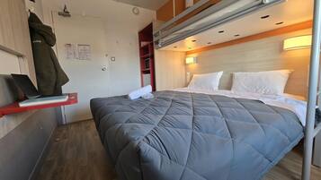 A compact hotel room with a bunk bed, a desk with a laptop and books, and a red shelf.