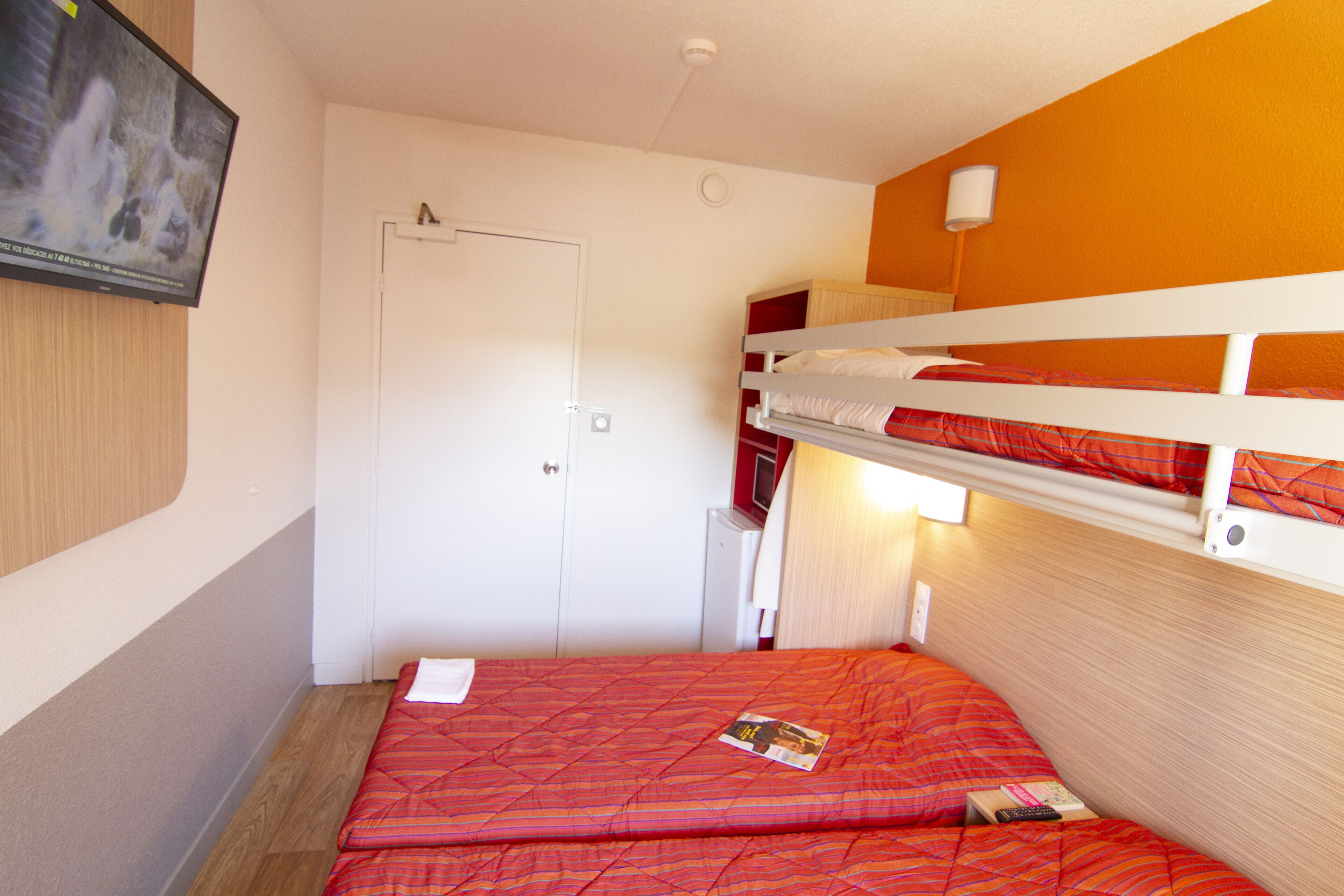 A room with two bunk beds, a television mounted on the wall, a door, and a small table with a remote control.
