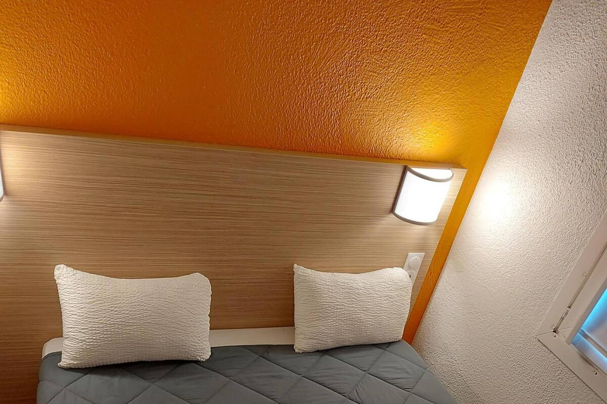 Un lit avec une couette grise matelassée, deux oreillers blancs et une tête de lit avec un revêtement en bois.
