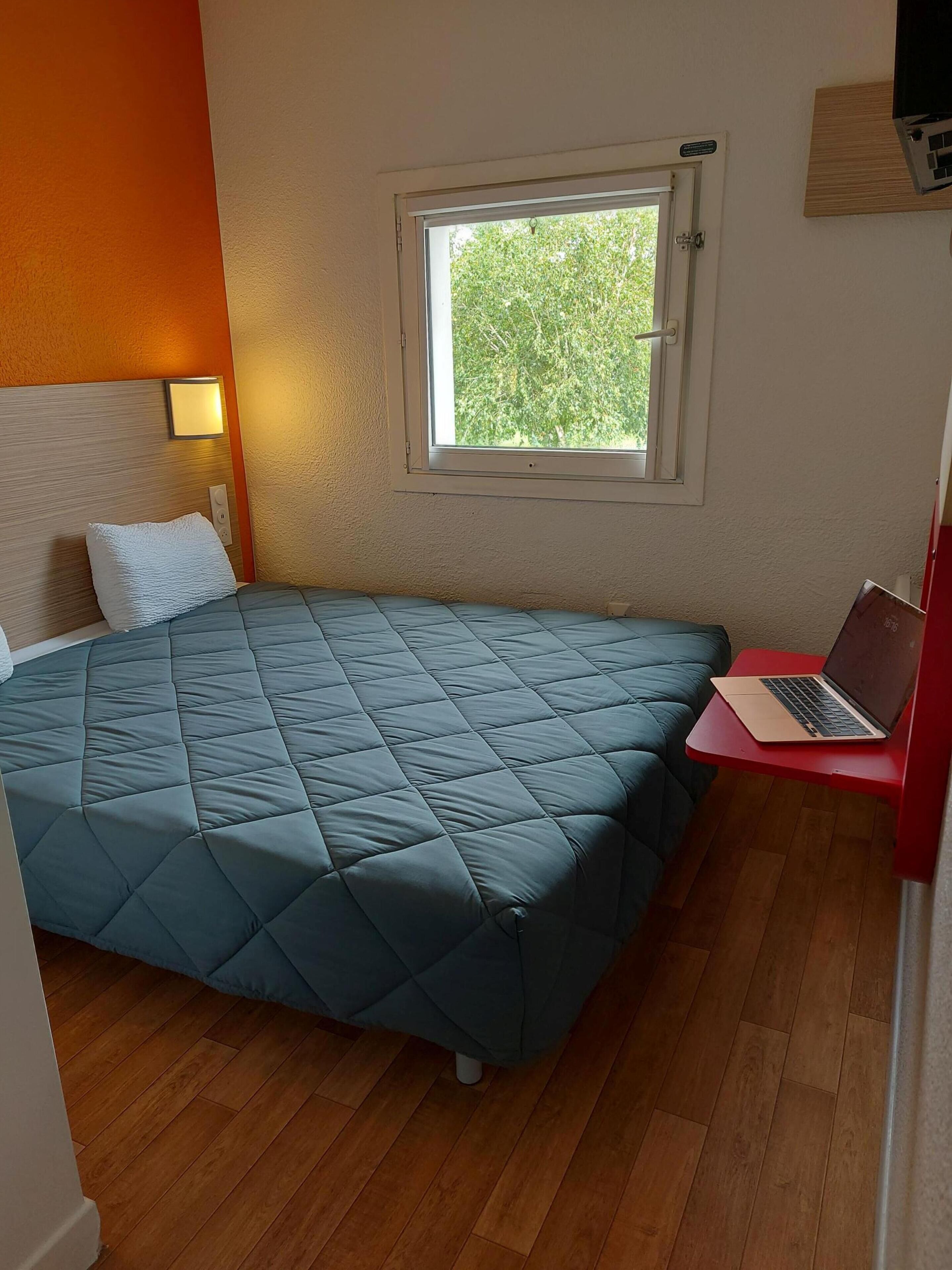 A room with a bed, a window, a laptop on a small table, and a lamp.