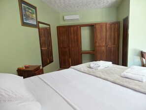 A bedroom with a bed, a wooden wardrobe, a small table with a red object, and a framed picture on the wall.