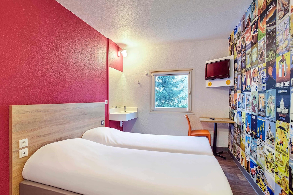 A hotel room with a bed, a desk, a TV, a window, and a wall decorated with travel posters.