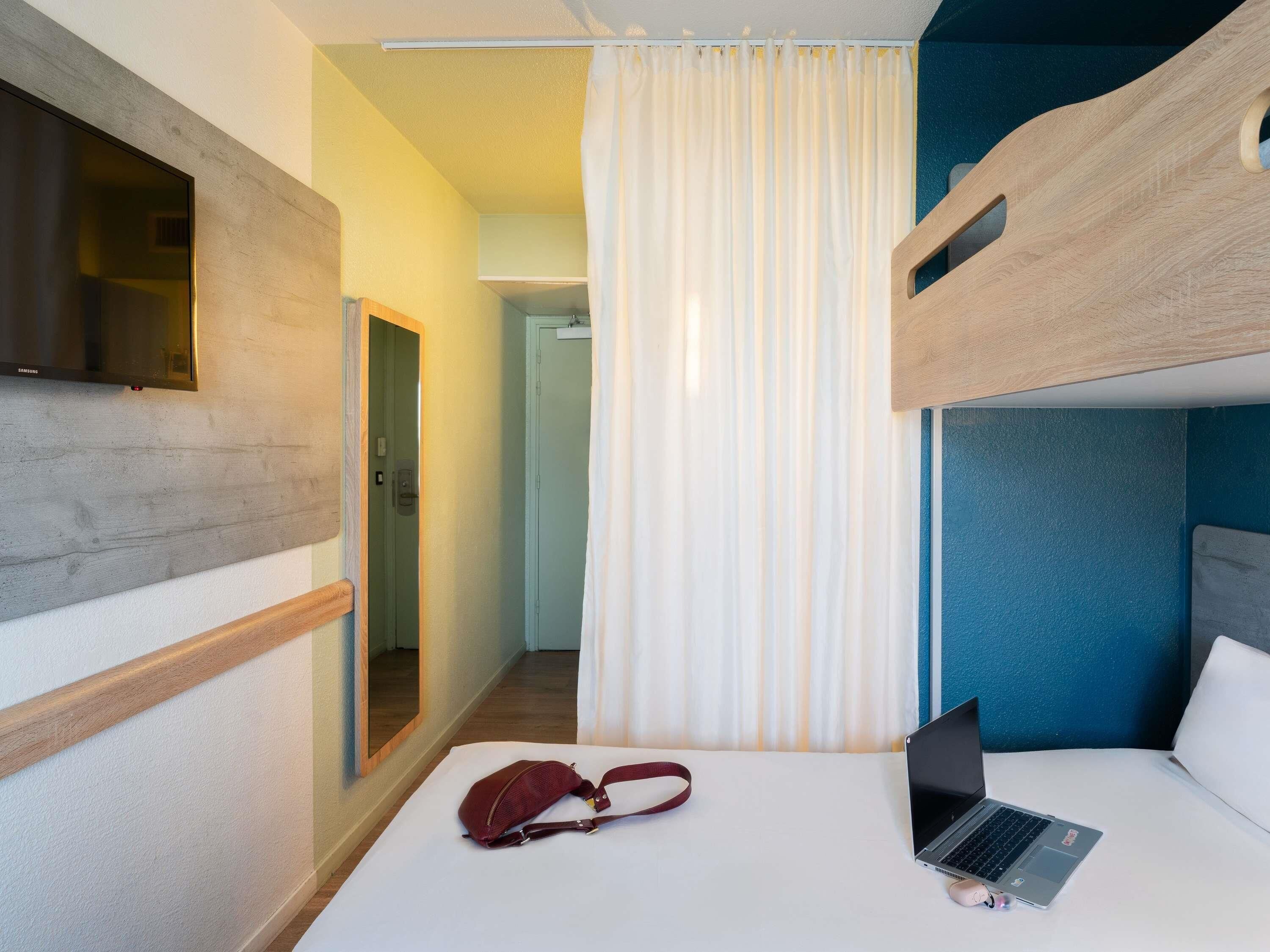 Une chambre d’hôtel avec un lit superposé, un ordinateur portable posé sur le lit, un sac rouge et un téléviseur fixé au mur.