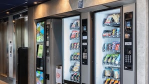 Een rij automaten met diverse snacks en drankjes, een prullenbak en een lift.