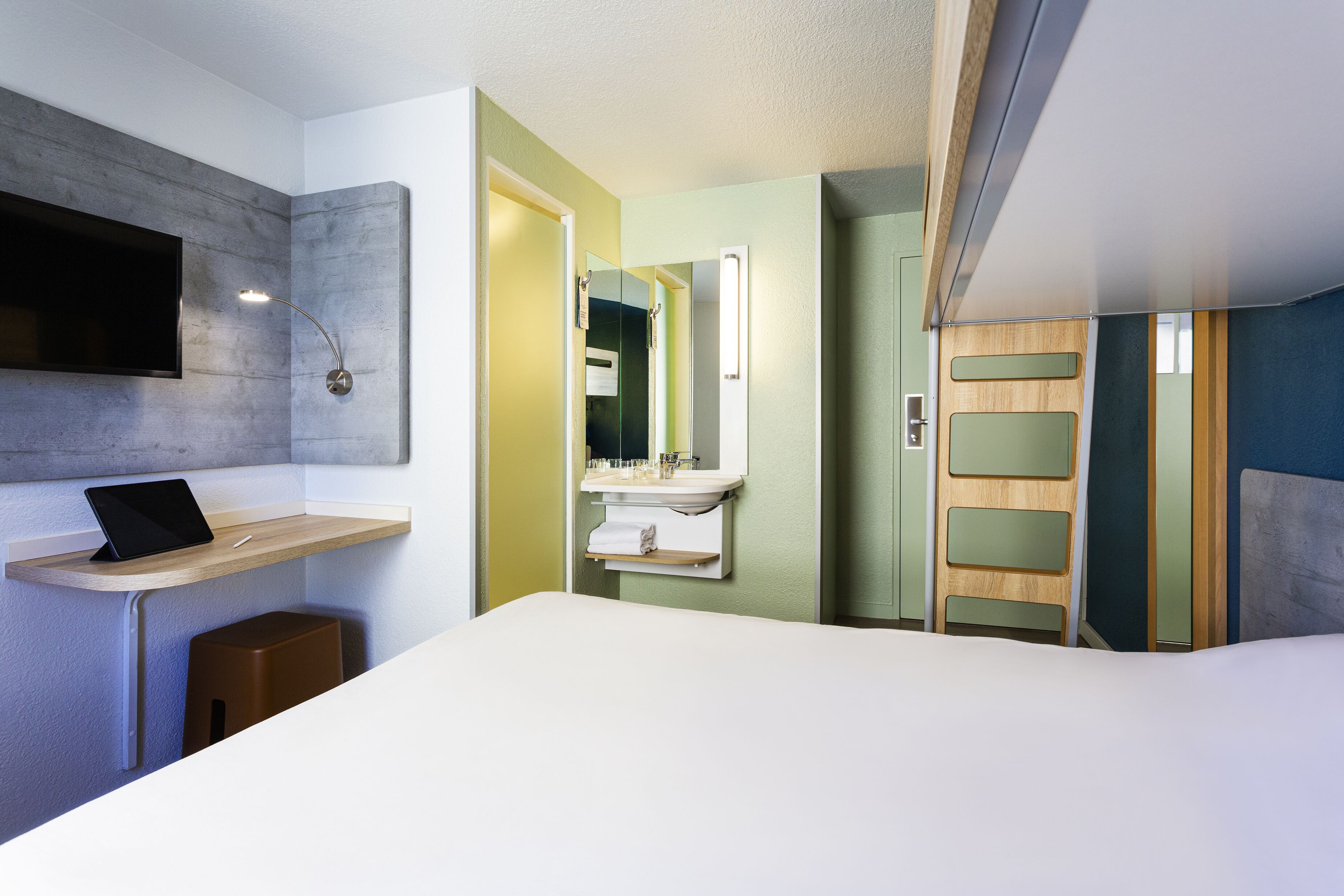 A modern hotel room with a bed, a desk with a laptop, a vanity with a mirror, and a wardrobe.
