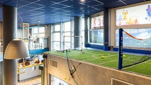 Een binnensportcomplex met een voetbalveld, een receptiebalie en een groot scherm waarop sportevenementen worden getoond.