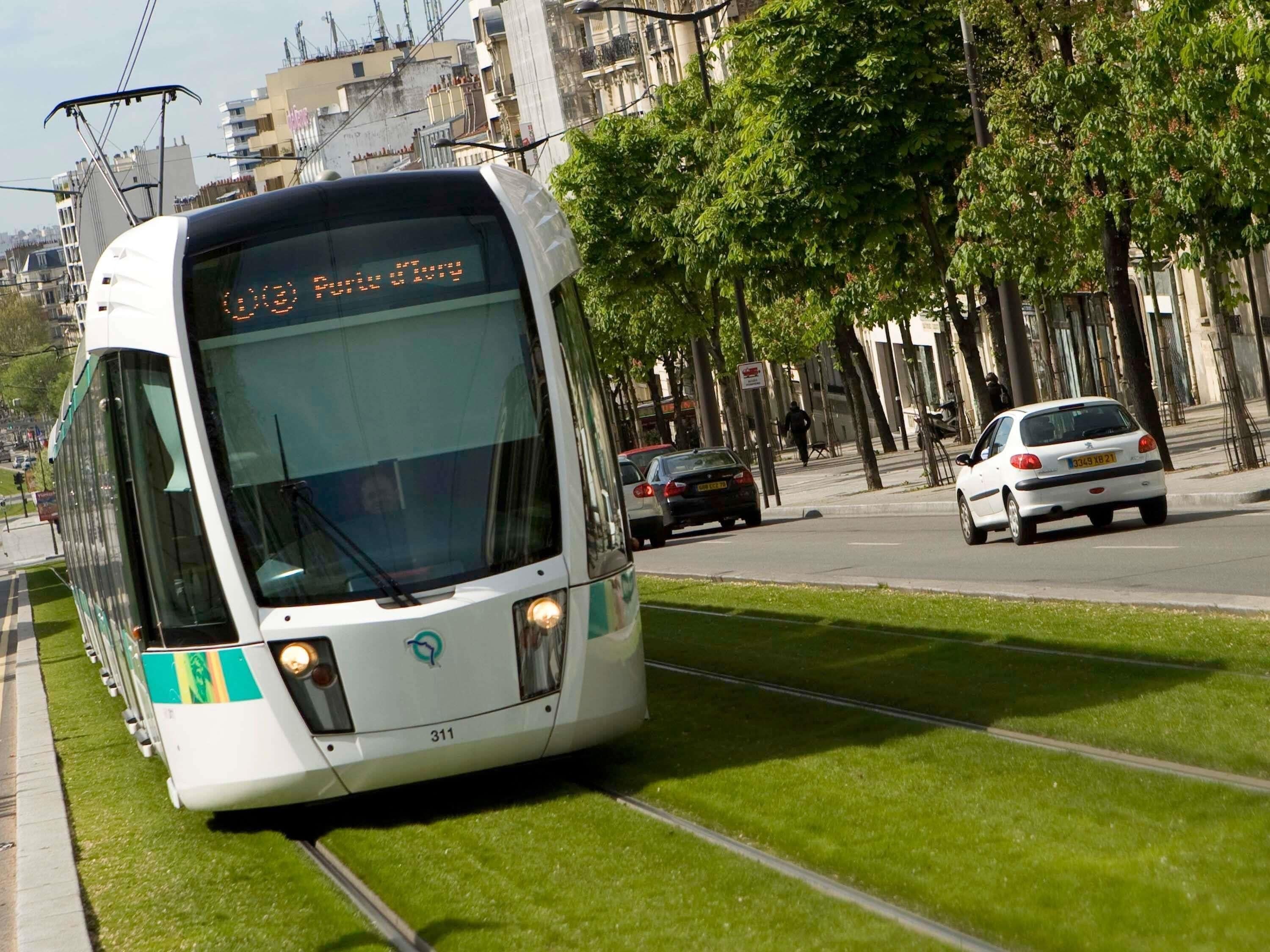 Un tramway circule sur des rails, équipé d’un écran numérique, et longe une route bordée de voitures et d’arbres.