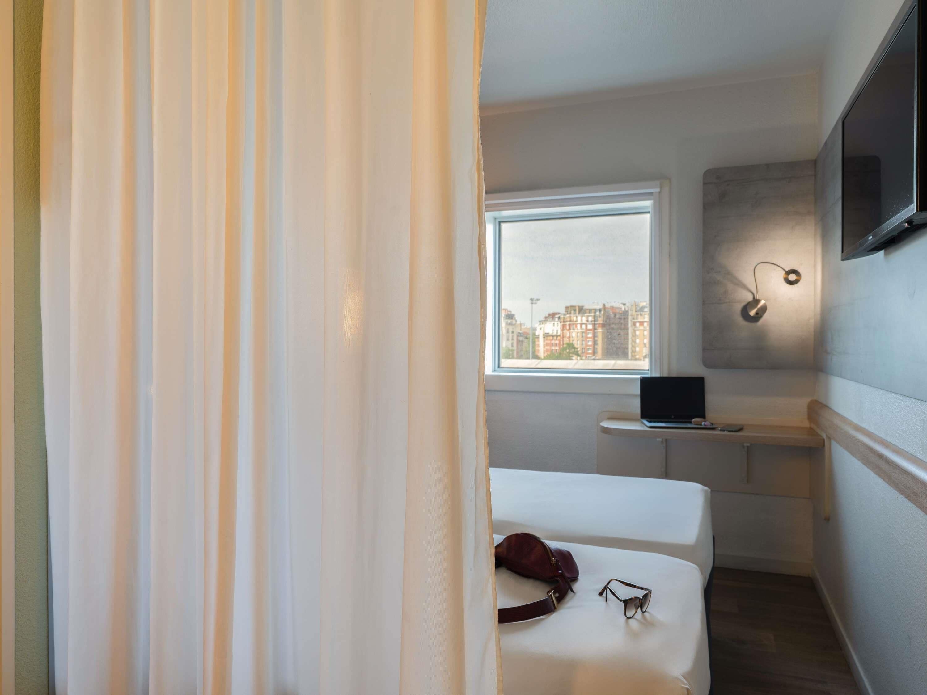 A hotel room with a bed, a small desk with a laptop, a window with a view of buildings, and a mounted TV.