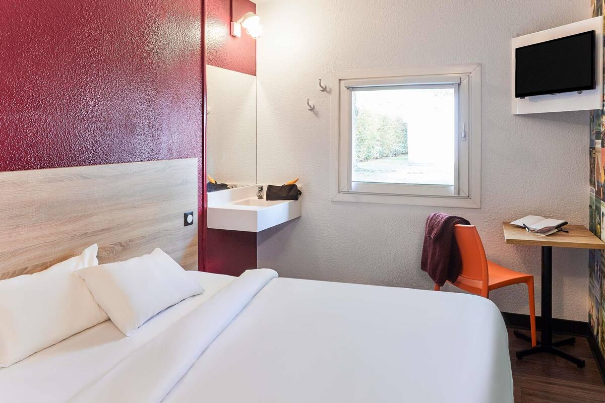 A hotel room with a bed, a desk with a chair, a sink, and a TV mounted on the wall.