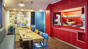 A compact dining area with a wooden table, blue chairs, and a bench. There are blue cabinets and a red counter with a coffee machine and snacks. - hotelF1 Clermont-Ferrand East (Clermont-Ferrand)