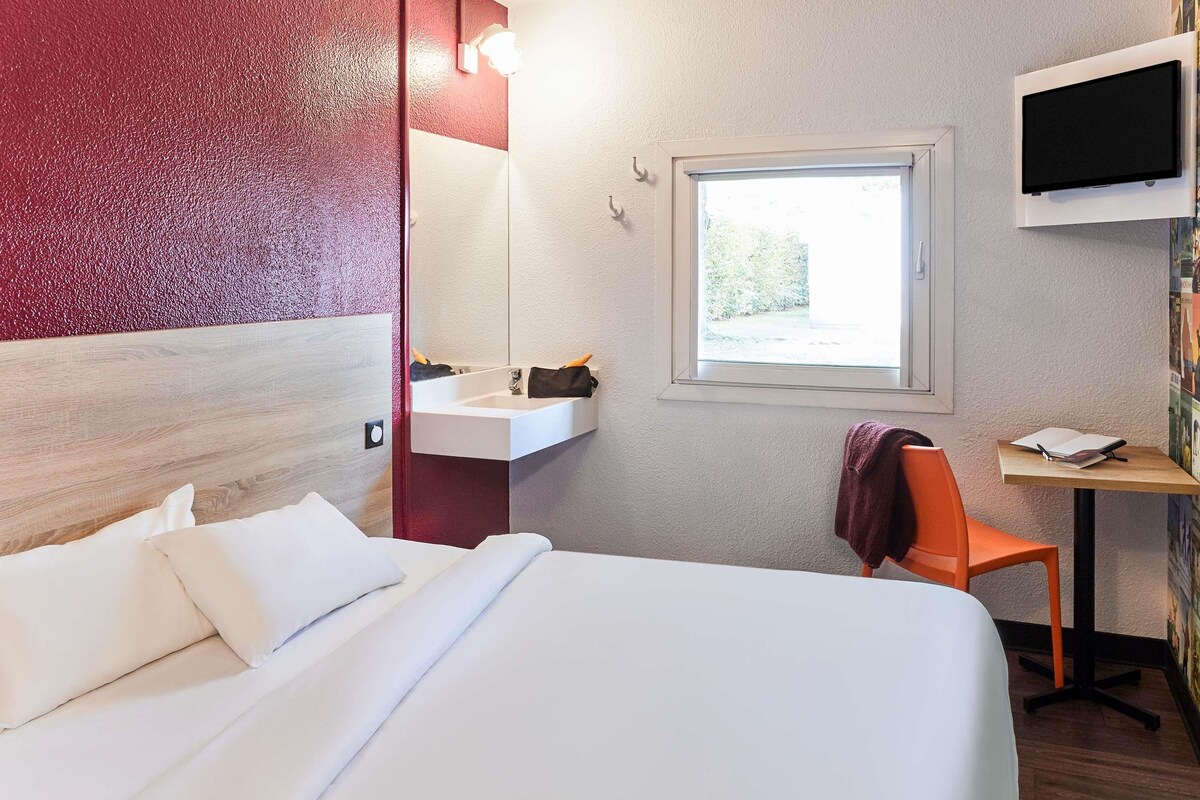 A hotel room with a bed, a desk with a chair, a sink, and a TV mounted on the wall.