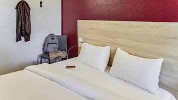 Una habitación de hotel con cama, almohadas blancas, cabecero de madera, pared de acento roja, perchero y una mochila en una silla.
