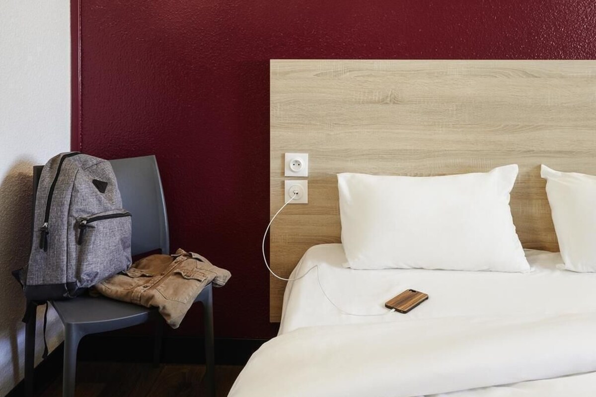 A hotel room with a wooden headboard, a gray chair with a backpack, and a white bed with pillows.