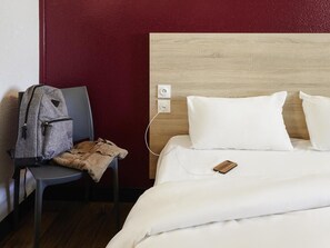 A hotel room with a wooden headboard, a gray chair with a backpack, and a white bed with pillows. - hotelF1 Clermont-Ferrand East (Clermont-Ferrand)
