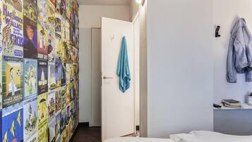 A hotel room with a bed, a desk with a lamp, a wall with travel posters, and a door with a towel hanging on it.