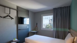 A hotel room with two beds, a TV mounted on the wall, a window with curtains, and a small table with a tennis racket and ball. - ibis budget Nice Aeroport Promenade des Anglais (Nice)