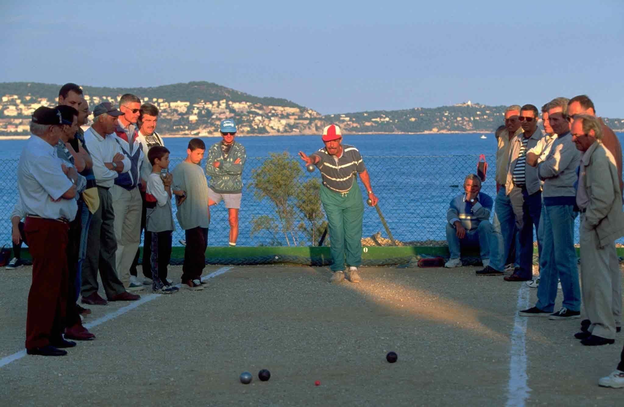 Un groupe de personnes joue à la pétanque sur un terrain aménagé, avec une vue pittoresque sur le bord de l’eau en arrière-plan.