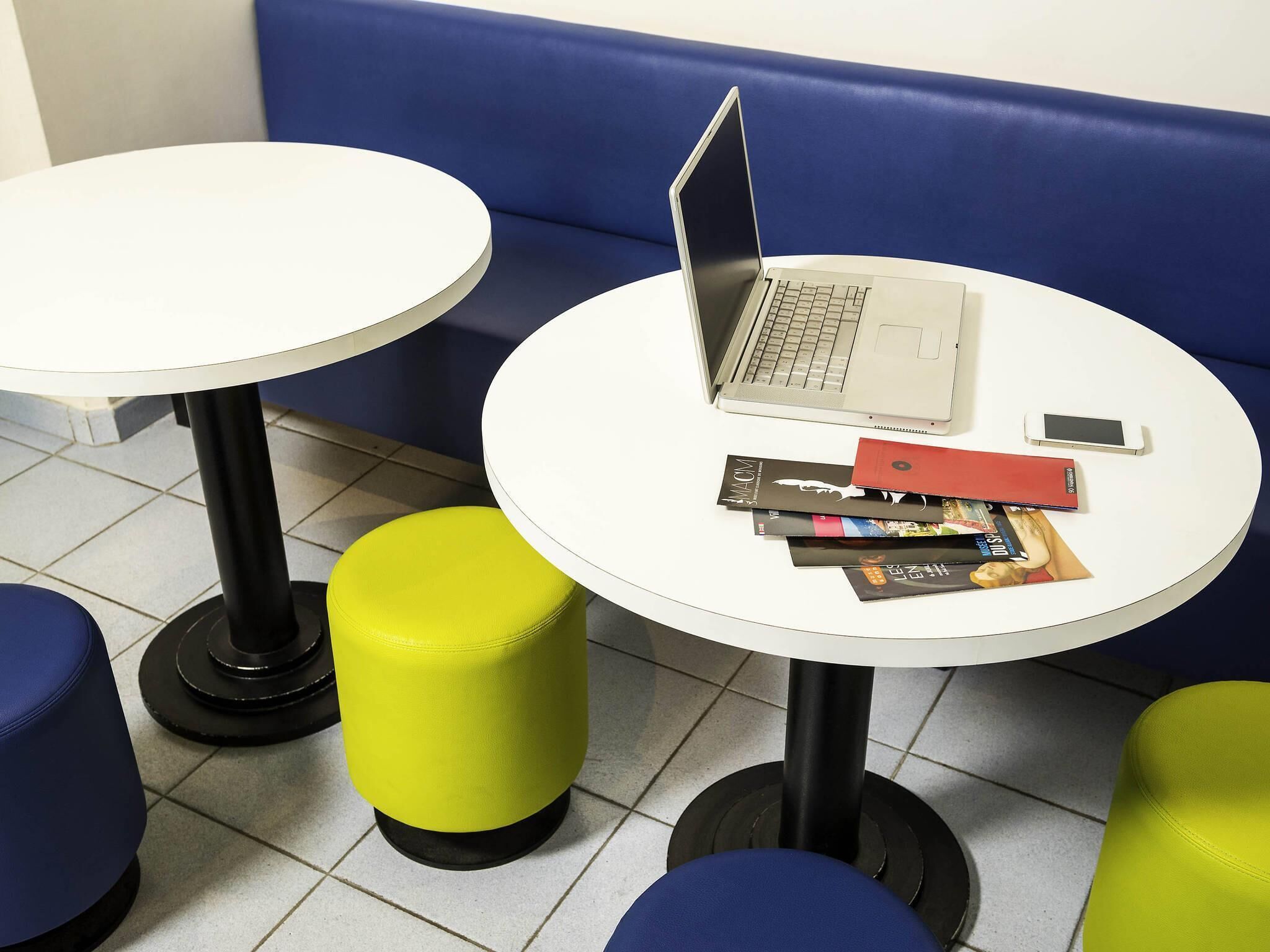 Uma área de estar com duas mesas redondas, uma com um laptop e livros, e dois banquinhos, um amarelo e outro azul.