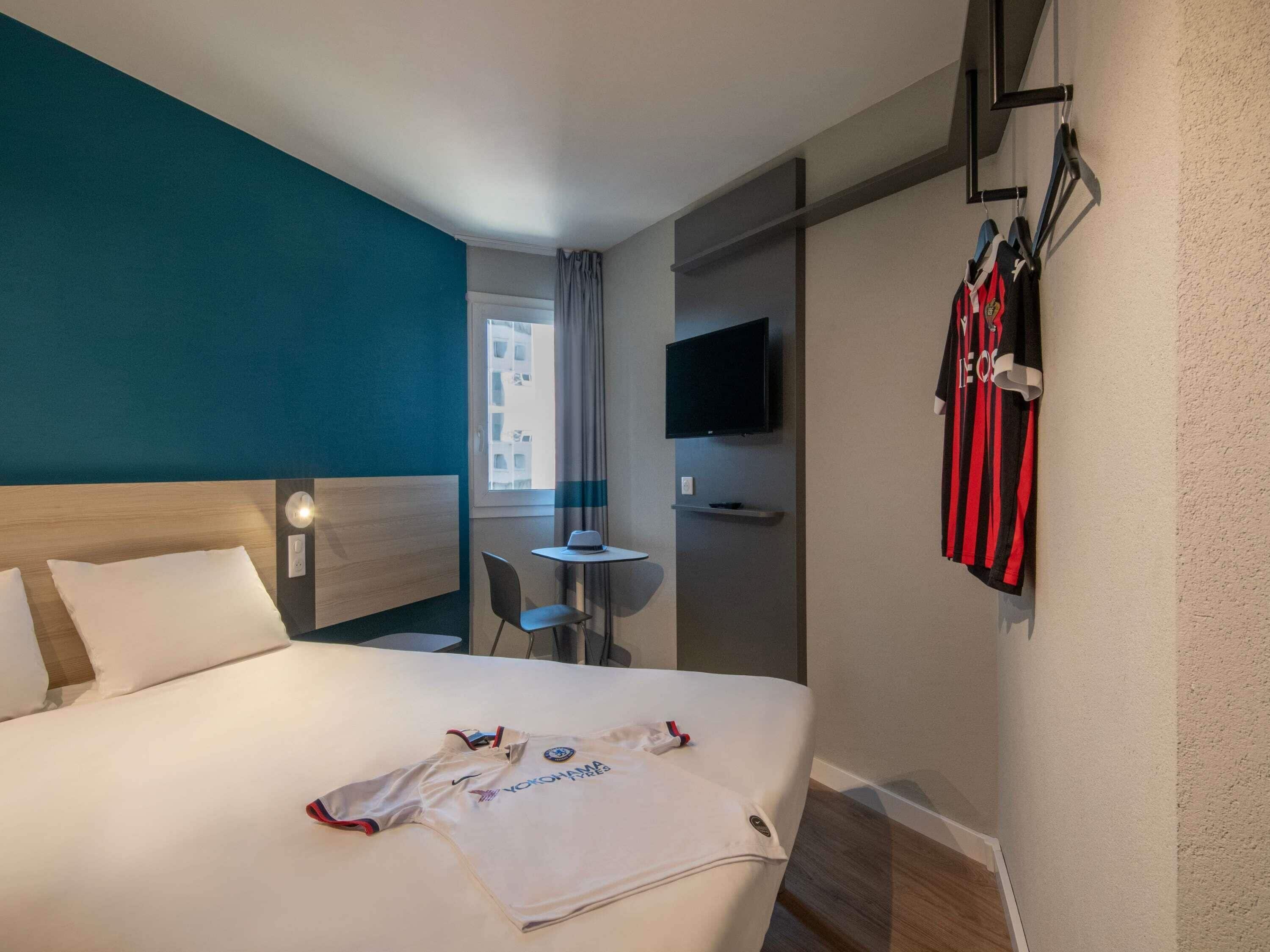 A hotel room with a bed, a TV mounted on the wall, a small table with a chair, and a window with curtains.