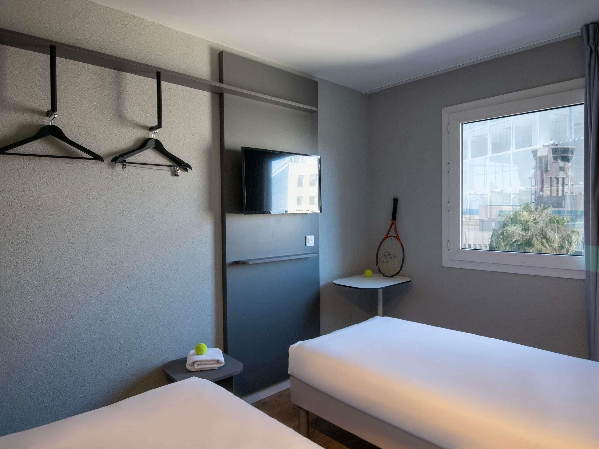 A hotel room with two beds, a TV mounted on the wall, a tennis racket, and a window with a view of buildings.