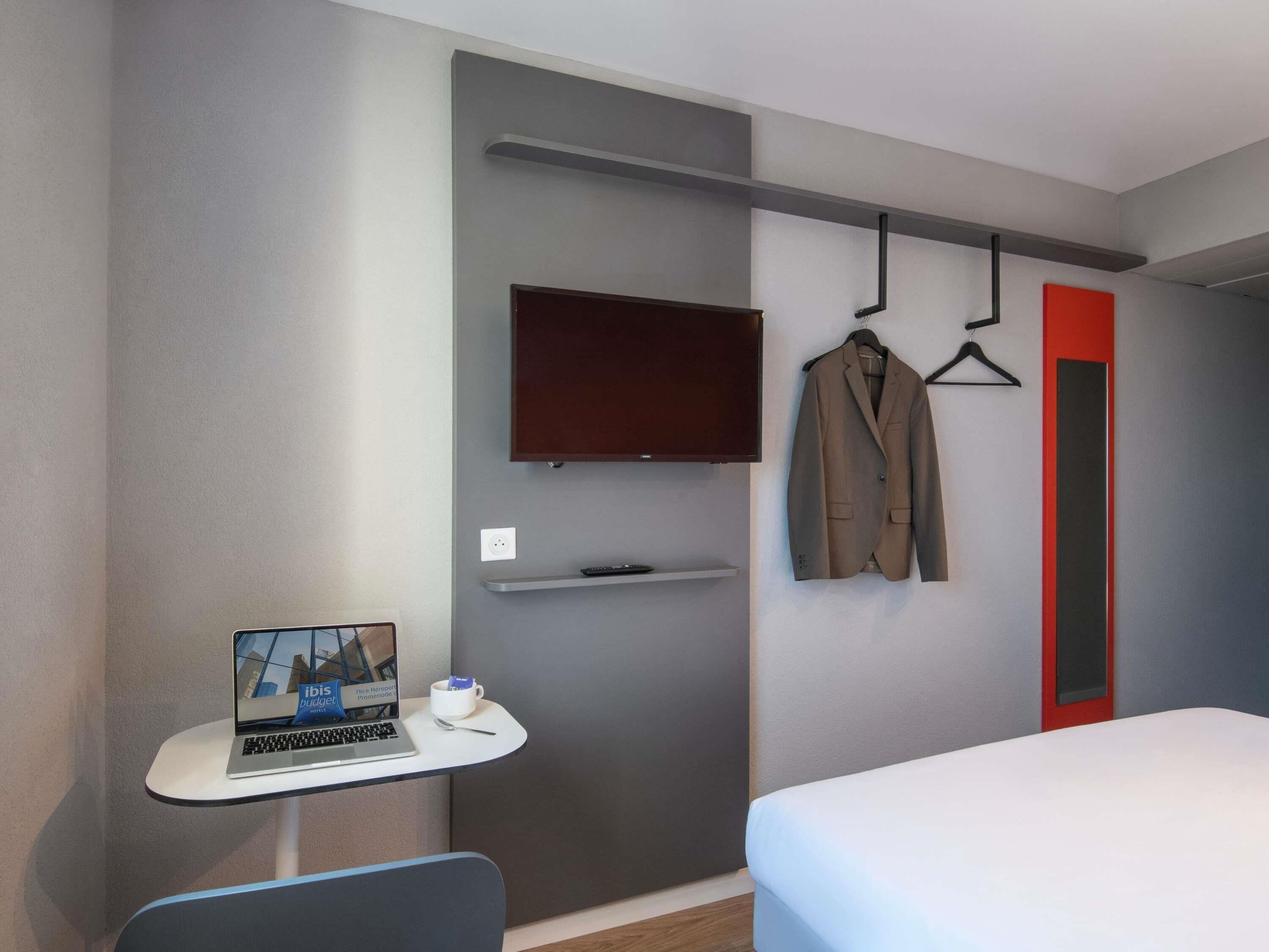A modern hotel room with a bed, a desk with a laptop, a TV mounted on the wall, and a coat hanger with a jacket.