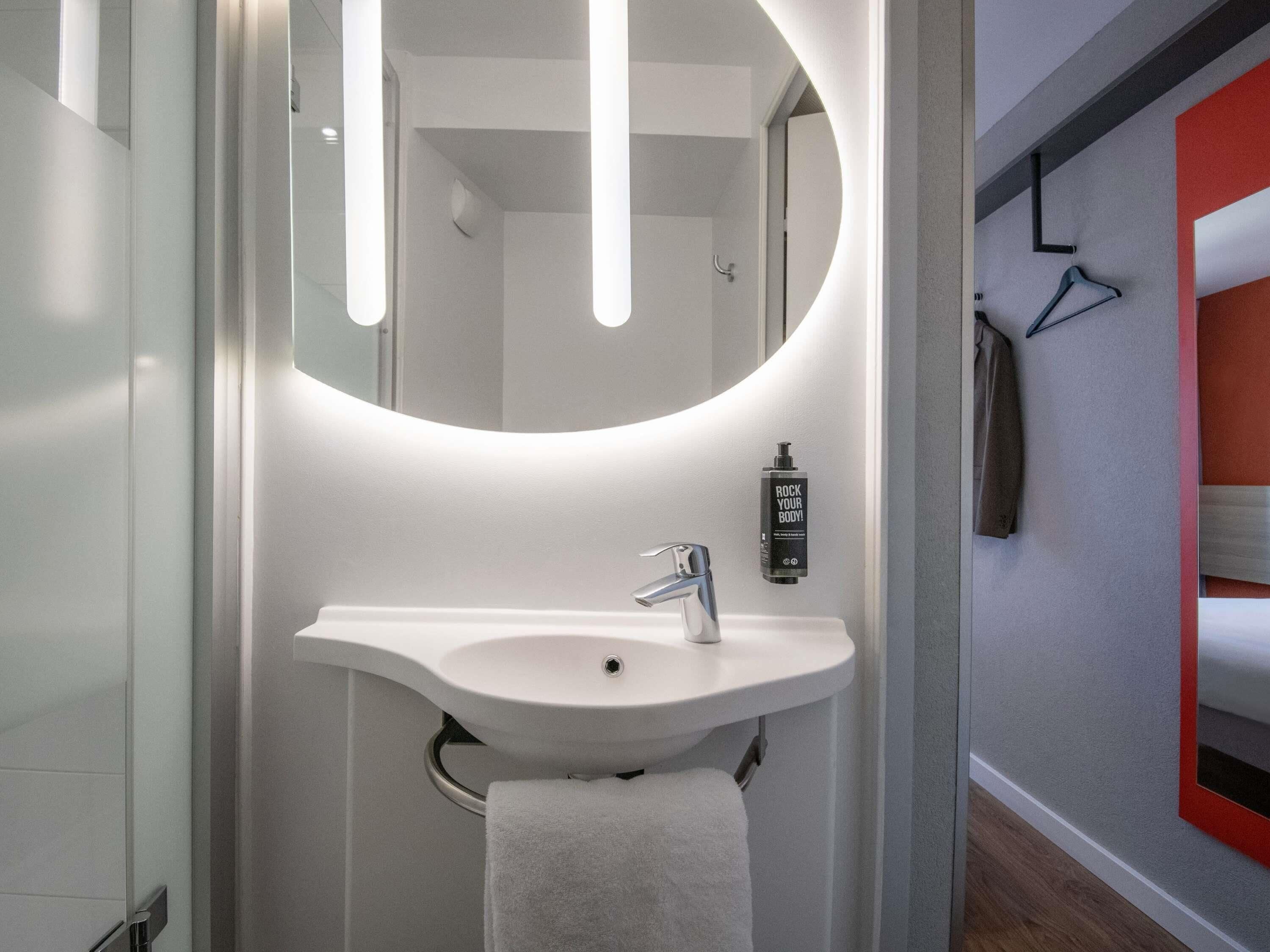 Un baño moderno con un lavamanos blanco, un espejo con luces verticales y una toalla colgada en un perchero.