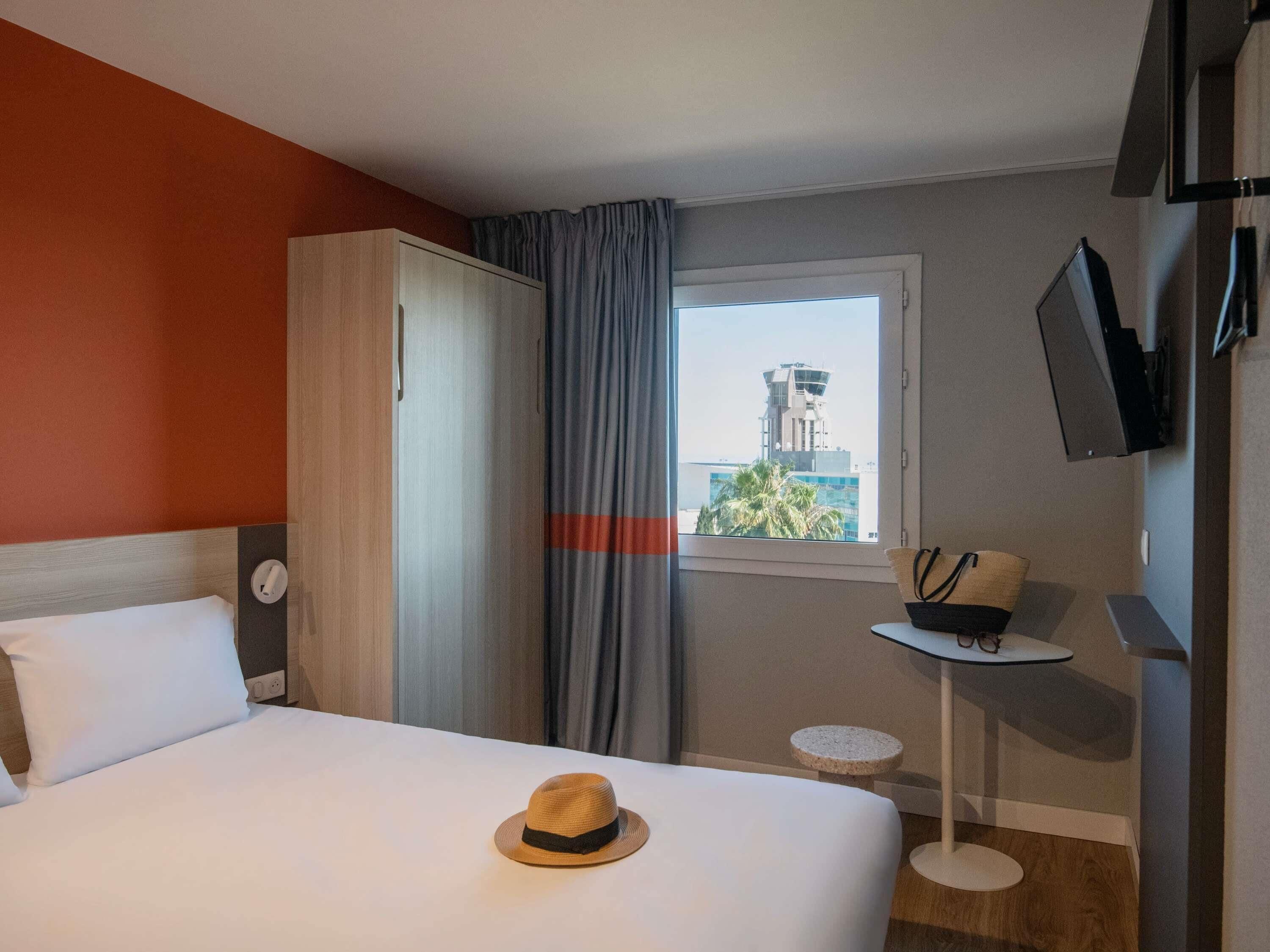 Una habitación de hotel con una cama, una mesita con un sombrero, un televisor y una ventana con vistas a un edificio.