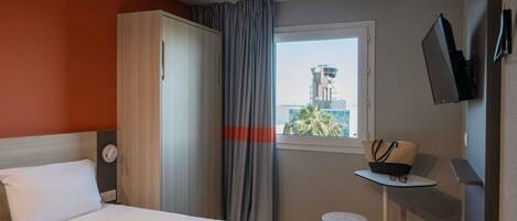 Una habitación de hotel con una cama, una mesita con un sombrero, un televisor y una ventana con vistas a un edificio.