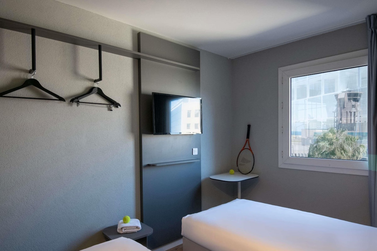 A hotel room with two beds, a TV, a tennis racket, and a window with a city view.
